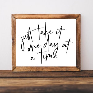 K&ouml;nnte beinhalten: Ein Schild mit Holzrahmen, wei&szlig;em Hintergrund und schwarzer Kalligraphie, auf dem "just take it one day at a time" steht.