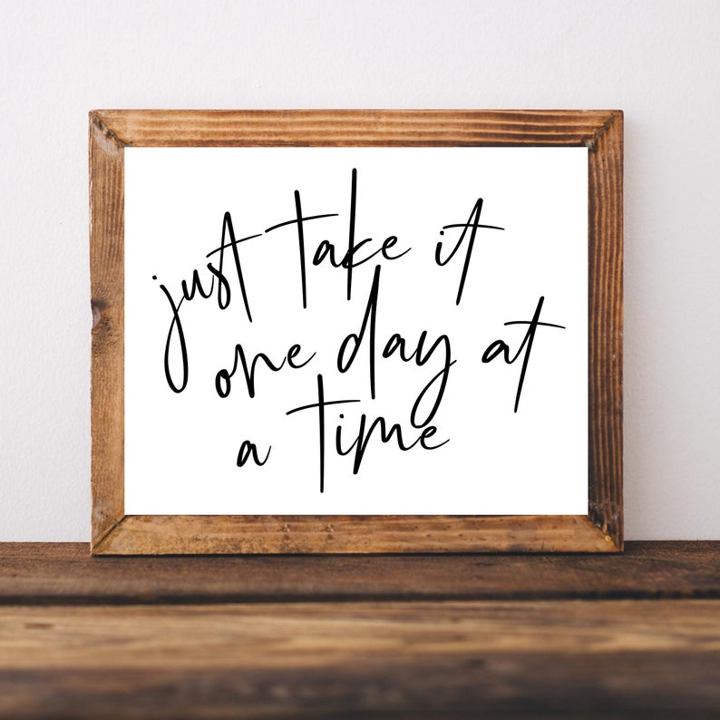 K&ouml;nnte beinhalten: Ein Schild mit Holzrahmen, wei&szlig;em Hintergrund und schwarzer Kalligraphie, auf dem "just take it one day at a time" steht.