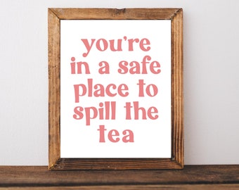 May include: A framed print with a white background and a pink text overlay that reads "you're in a safe place to spill the tea". The frame is made of brown wood.