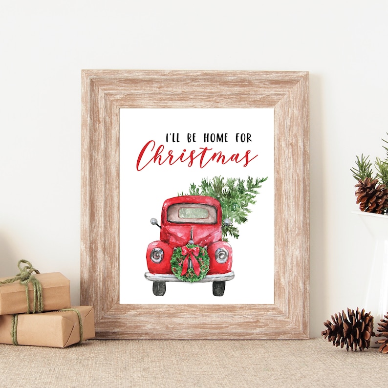 Puede incluir: Un cami&oacute;n vintage rojo con un &aacute;rbol de Navidad en la parte trasera y una corona en la parte delantera. El texto "I'll be home for Christmas" est&aacute; escrito en rojo sobre el cami&oacute;n.