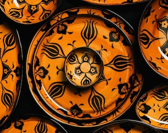 May include: A collection of ceramic plates and a small bowl featuring a vibrant orange background with intricate black floral and tulip-inspired patterns. The circular dinnerware is arranged in a top-down view, showcasing consistent motifs across all pieces.