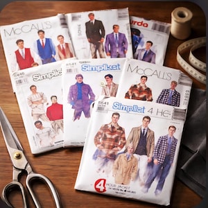 May include: A collection of vintage sewing patterns for men's clothing, including jackets, shirts, and vests. The patterns are from brands like McCall's and Simplicity, with images of the finished garments on the covers. A pair of scissors, measuring tape, and thread spool are also visible.