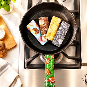 May include: A collection of five rectangular fabric skillet handle covers featuring various prints including donuts, coffee cups, bread, text, and footballs. The covers are quilted and designed to fit over the handle of a black cast iron skillet.