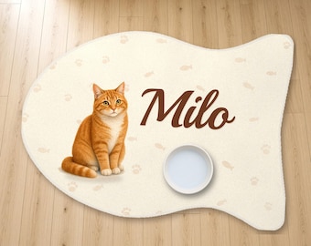 May include: A fish-shaped cream-coloured pet feeding mat featuring a printed orange tabby cat, the name Milo in brown script, and a pattern of small fish and paw prints. A plain white circular bowl sits on the mat surface.