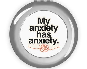 May include: A round, silver-colored compact mirror with a white center insert. The text "My anxiety has anxiety." is printed in black, with a small, red, tangled line graphic positioned directly underneath the text.