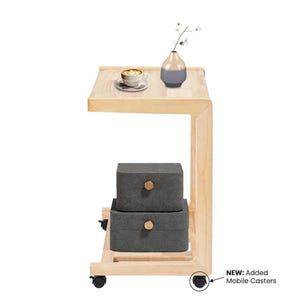 May include: A light-colored wooden C-shaped side table with mobile casters. The table features a flat top surface holding a coffee cup on a saucer and a small vase, with two stacked dark gray storage boxes with gold knobs on the lower shelf.