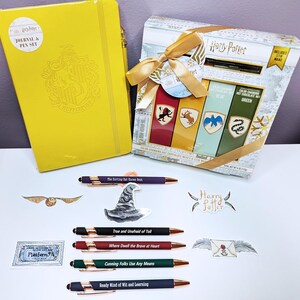 May include: A yellow Harry Potter journal and pen set next to a gift box of color-changing hot chocolate mix. The box is tied with a gold ribbon. Also included are several pens with quotes and stickers with Harry Potter imagery.