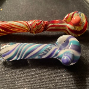 Puede incluir: Dos pipas para fumar de vidrio con patrones en espiral. Una pipa es roja, blanca y dorada, y la otra es azul, blanca y morada.