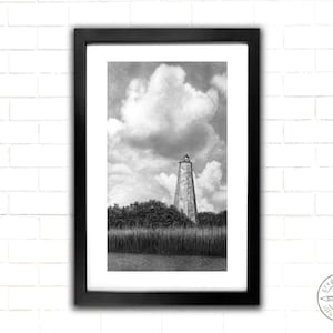 May include: A black and white photograph of a lighthouse with a large cloud in the sky. The lighthouse is tall and narrow, with a white tower and a black roof. The cloud is fluffy and white, and it covers most of the sky. The lighthouse is surrounded by trees and grass.
