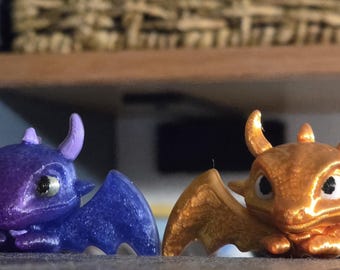 May include: Two small dragon figurines, one in a shimmering deep purple and the other in a metallic gold finish. Both creatures feature pointed horns, large dark eyes, and outstretched wings, crafted with a textured, layered appearance.
