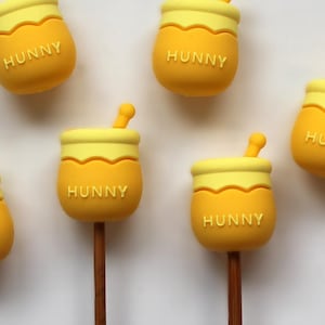 May include: Six yellow honey pot shaped silicone toppers with the word "Hunny" on each. The toppers are on wooden sticks.