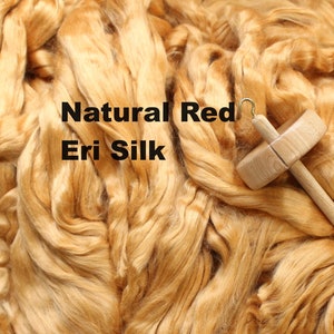 May include: A large pile of natural red eri silk fiber. The silk is a light orange color and is arranged in a loose, fluffy pile. A wooden spinning wheel is lying on top of the silk.