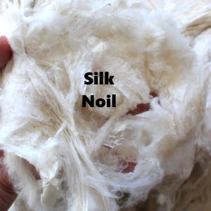May include: Close-up of a handful of off-white silk noil fibers. The fibers are soft and textured, with the text "Silk Noil" in black. The fibers are a natural, creamy color, and appear to be loosely packed.