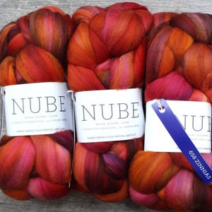 May include: Four bundles of Nube merino wool roving in a vibrant blend of red, orange, and purple hues. Each bundle is labeled with a white tag and a blue tag that reads "658 Zinnias". The wool is arranged on a wooden surface.