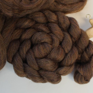 May include: A close-up of brown wool roving and a wooden drop spindle. The wool is a rich, dark brown color, with a soft, textured appearance. The drop spindle is made of light-colored wood and has a simple, functional design. The wool is arranged in a circular pattern.