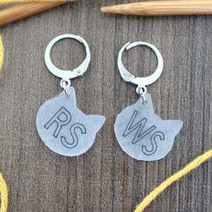 May include: Two silver cat-shaped charms with the letters "RS" and "WS" printed on them. The charms are hanging from silver hoops.