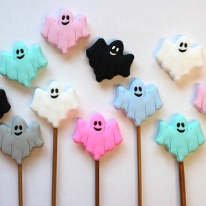 May include: A set of 12 ghost-shaped cake toppers in various colors, including pink, blue, gray, white, and black. Each ghost has a friendly face and is attached to a wooden stick.