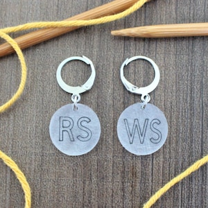 May include: Two silver knitting stitch markers with clear round tags. The tags are labeled "RS" and "WS".