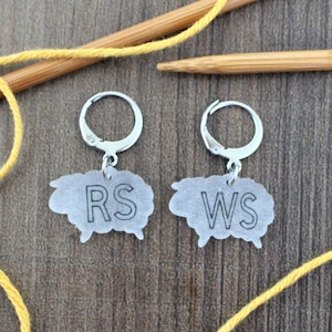 May include: Two silver sheep charms with hoop attachments. The charms are labeled "RS" and "WS" for right side and wrong side of knitting.