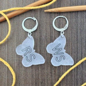 May include: Two silver butterfly-shaped stitch markers with the words "RS" and "WS" printed on them. The markers are hanging from silver hoops.