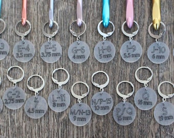May include: A set of 15 crochet hooks with clear plastic size indicators. The hooks are arranged in two rows on a wooden surface. The size indicators include letters and millimeters. The hooks are in various colors, including blue, pink, green, and gold.