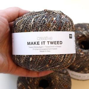 May include: A close-up of three balls of tweed yarn. The yarn is a mix of brown, black, and white flecks. The label on the front ball reads "MAKE IT TWEED". The yarn is made in Italy.