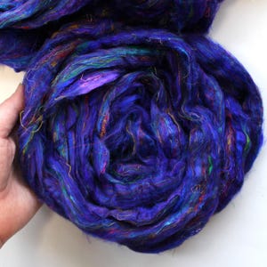 May include: A large, tightly wound skein of vibrant blue yarn with streaks of teal, purple, and gold. The yarn appears to be silk or a similar fiber, with a soft, textured appearance. The skein is held in a hand against a white background.