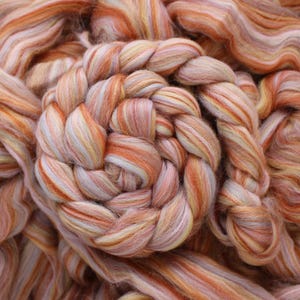May include: Close-up of a pile of colorful fiber for crafting. The fiber is a mix of orange, peach, yellow, and white, with a braided section in the center. The texture appears soft and fluffy, ideal for spinning or felting projects.