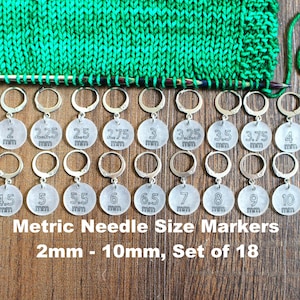 May include: Set of 18 silver knitting needle size markers, with numbers 2mm to 10mm printed on each marker. The markers are arranged in two rows on a wooden surface.
