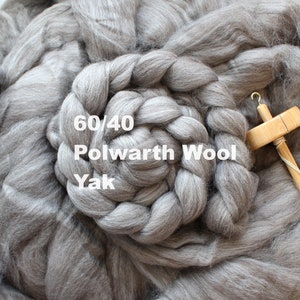 May include: A close-up of a gray 60/40 Polwarth Wool Yak fiber blend. The fiber is arranged in a braided circle and a wooden spinning wheel is visible in the background.