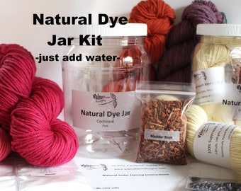 May include: Natural dye kit with four glass jars containing different dye ingredients: cochineal, madder root, logwood, and a jar of alum crystals. The kit also includes three skeins of yarn in various shades of pink and white.