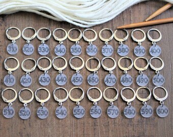 May include: A set of 30 silver stitch markers with clear plastic circles numbered from 310 to 600. The markers are arranged in three rows of ten on a wooden surface.