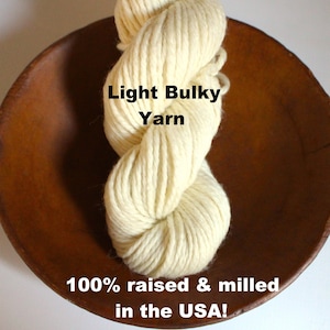 May include: A skein of light bulky yarn, 100% raised and milled in the USA. The yarn is a creamy white color and is displayed in a wooden bowl.