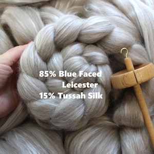 May include: A close-up of a braid of light gray wool fiber with a spinning wheel in the background. The wool is labeled "85% Blue Faced Leicester 15% Tussah Silk".