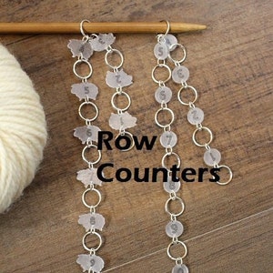 May include: Three silver chains with clear plastic row counters hanging from a wooden dowel. Each counter has a black number printed on it, starting at 1 and going up to 10. The text "Row Counters" is printed on the wooden dowel.