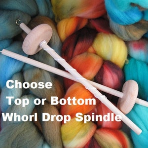 May include: Two wooden drop spindles with a whorl on each end. The spindles are crossed over each other. The spindles are surrounded by colorful yarn. The text "Choose Top or Bottom Whorl Drop Spindle" is written below the spindles.