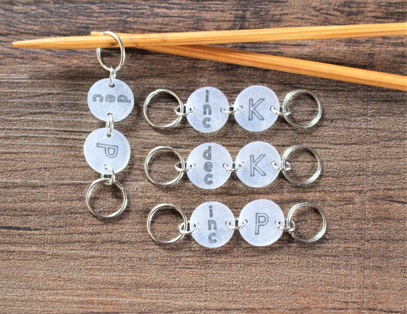 May include: Set of six silver knitting stitch markers with clear plastic discs. The discs have the words "inc", "dec", "k", "p", "co", and "inc" printed on them.