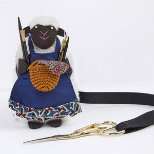 May include: A black and white sheep plush toy wearing a blue and white apron with a brown crocheted basket. The sheep is holding knitting needles and a pair of gold scissors are attached to a black ribbon.