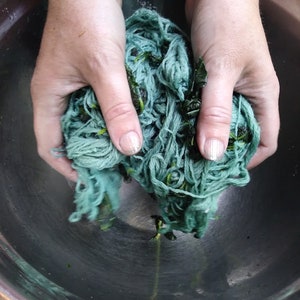 May include: A person's hands are squeezing a ball of teal blue yarn with green leaves mixed in. The yarn is in a stainless steel bowl.