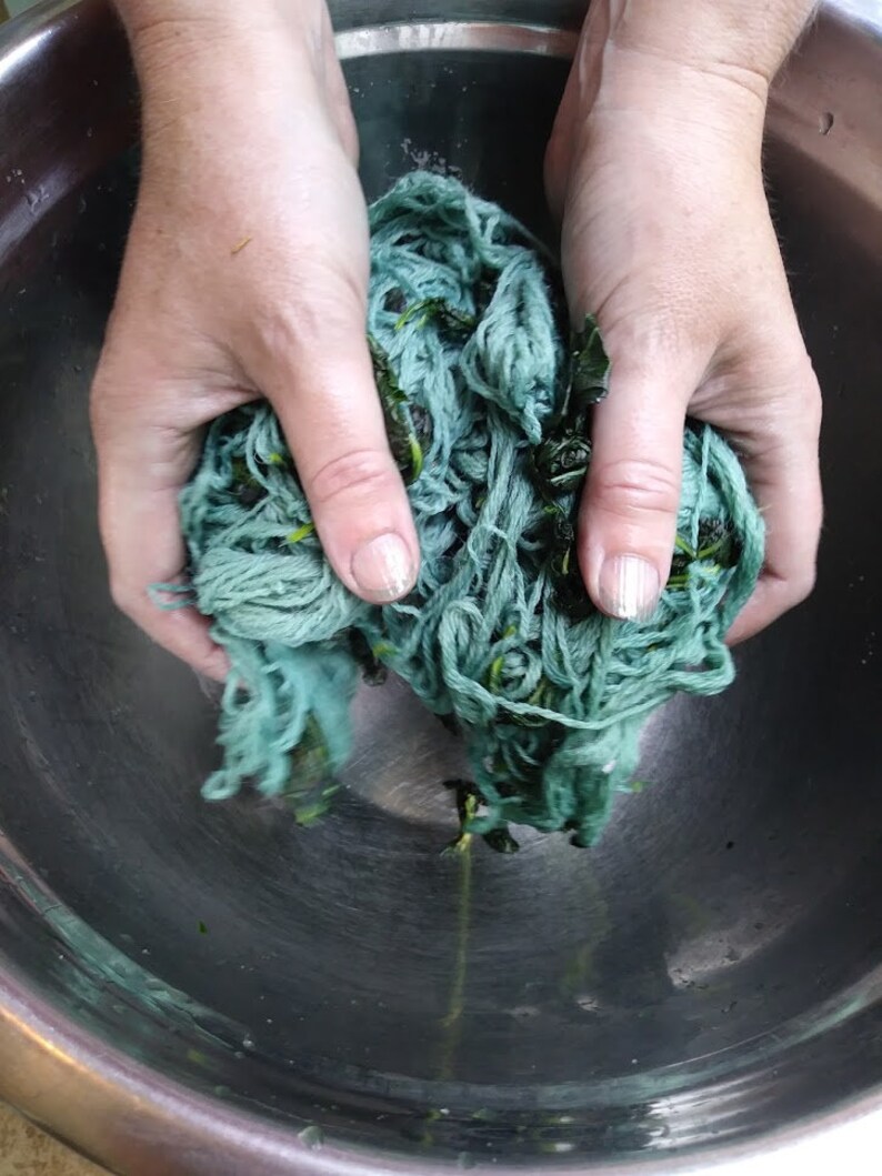 May include: A person's hands are squeezing a ball of teal blue yarn with green leaves mixed in. The yarn is in a stainless steel bowl.