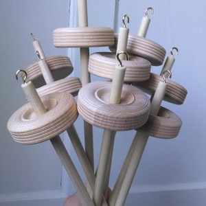 May include: A group of seven wooden spinning wheels with gold hooks. The wheels are stacked on top of each other and are held in a hand.