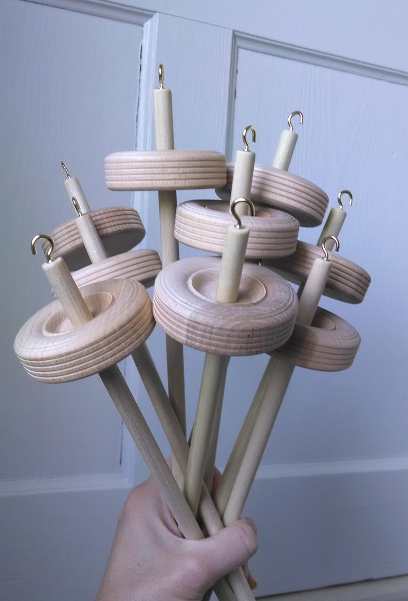 May include: A group of seven wooden spinning wheels with gold hooks. The wheels are stacked on top of each other and are held in a hand.