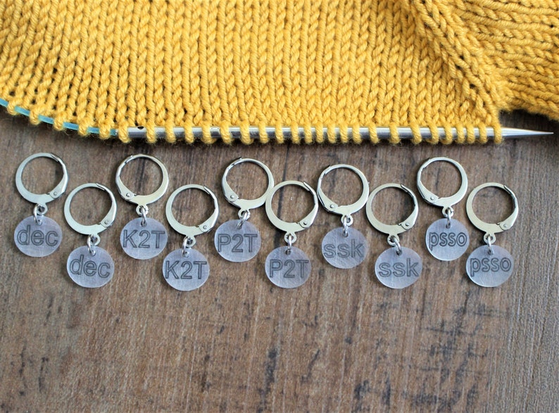 May include: Eight clear plastic stitch markers with metal rings. The markers are labeled with knitting abbreviations: dec, K2T, P2T, ssk, psso.