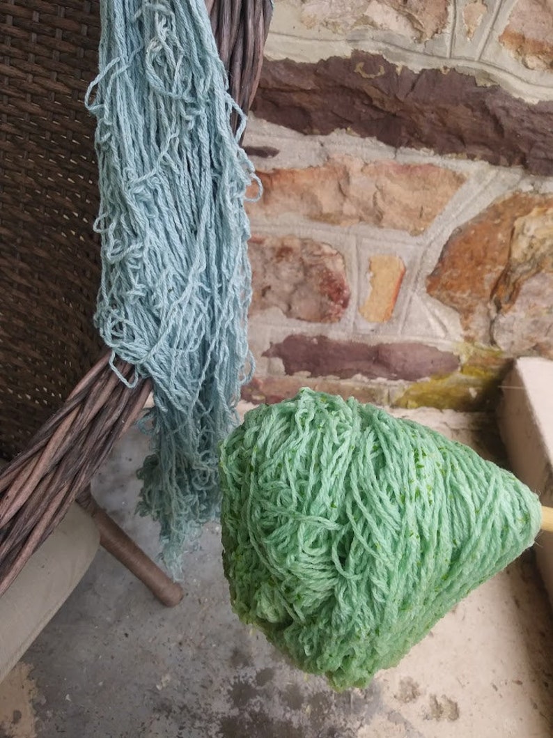 May include: Two skeins of yarn, one light blue and one green, are hanging from a wicker basket. The yarn is soft and fluffy, and it would be perfect for knitting or crocheting.