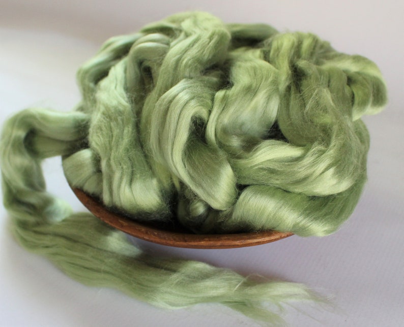 Puede incluir: Un bol lleno de una gran cantidad de fibra de seda verde claro. La fibra es suave y esponjosa, y es perfecta para hilar o fieltro.
