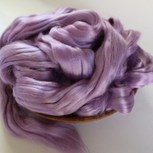 Peut inclure: Un tas de fibres de soie de couleur lavande, dens&eacute;ment tass&eacute;es dans un bol en bois. Les fibres ont un &eacute;clat doux et soyeux, sugg&eacute;rant un mat&eacute;riau de haute qualit&eacute;. L'image est bien &eacute;clair&eacute;e, mettant en valeur la texture et la couleur de la soie.