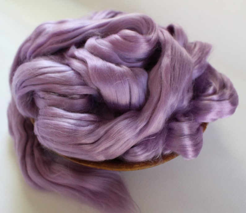 Peut inclure: Un tas de fibres de soie de couleur lavande, dens&eacute;ment tass&eacute;es dans un bol en bois. Les fibres ont un &eacute;clat doux et soyeux, sugg&eacute;rant un mat&eacute;riau de haute qualit&eacute;. L'image est bien &eacute;clair&eacute;e, mettant en valeur la texture et la couleur de la soie.