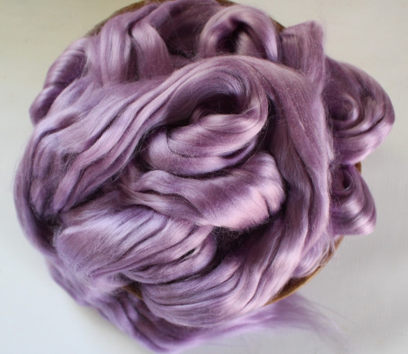 Peut inclure: Un tas de fibres douces de couleur lavande, peut-&ecirc;tre de la soie ou de la laine, remplit un panier rond et peu profond. Les fibres ont un &eacute;clat subtil, avec des reflets et des ombres qui cr&eacute;ent de la profondeur. La texture semble lisse et luxueuse, sugg&eacute;rant un mat&eacute;riau artisanal.