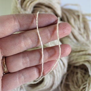 K&ouml;nnte beinhalten: Nahaufnahme einer Hand, die zwei F&auml;den aus Naturfasergarn h&auml;lt. Das Garn ist hellbeige und hat eine leicht raue Textur.