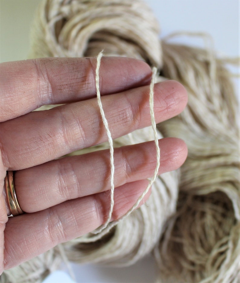 K&ouml;nnte beinhalten: Nahaufnahme einer Hand, die zwei F&auml;den aus Naturfasergarn h&auml;lt. Das Garn ist hellbeige und hat eine leicht raue Textur.